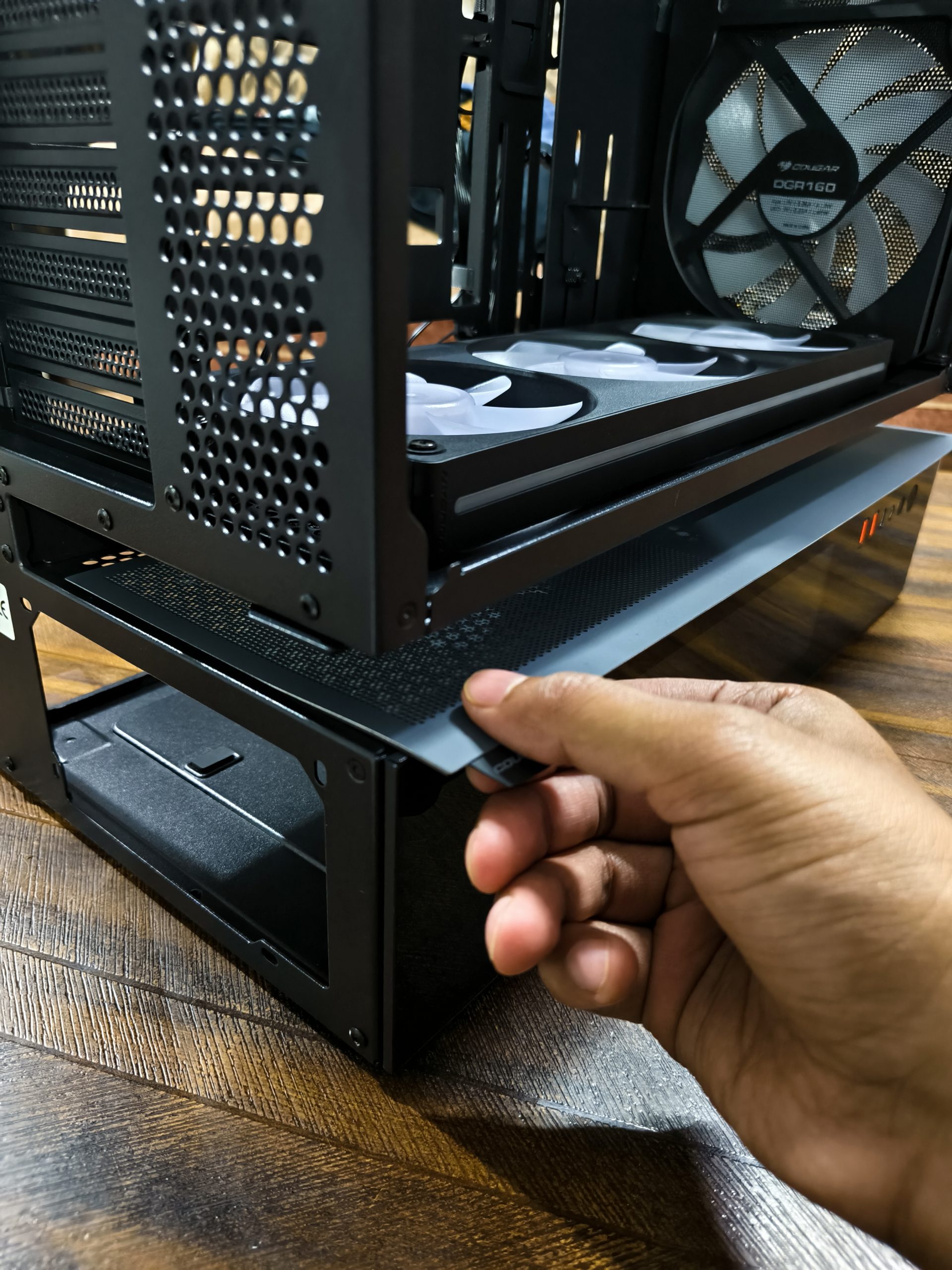 Ένα άτομο συναρμολογεί μια θήκη υπολογιστή, εγκαθιστά έναν ανεμιστήρα Cougar DQR160 σε ένα ξύλινο γραφείο.
