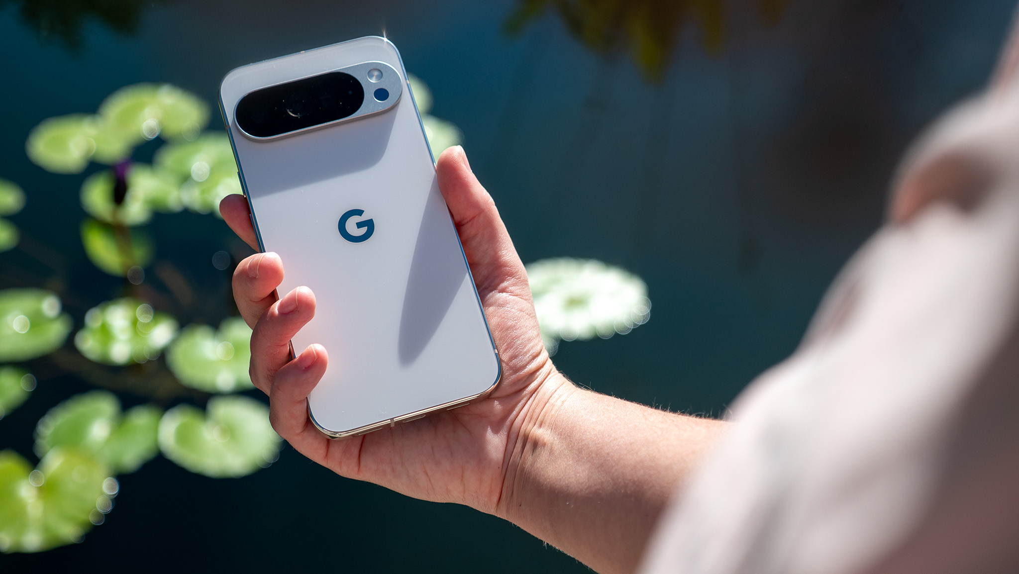 Κρατώντας ένα λευκό Google Pixel 10 Pro XL μπροστά από μια λίμνη γεμάτη με κρίνους