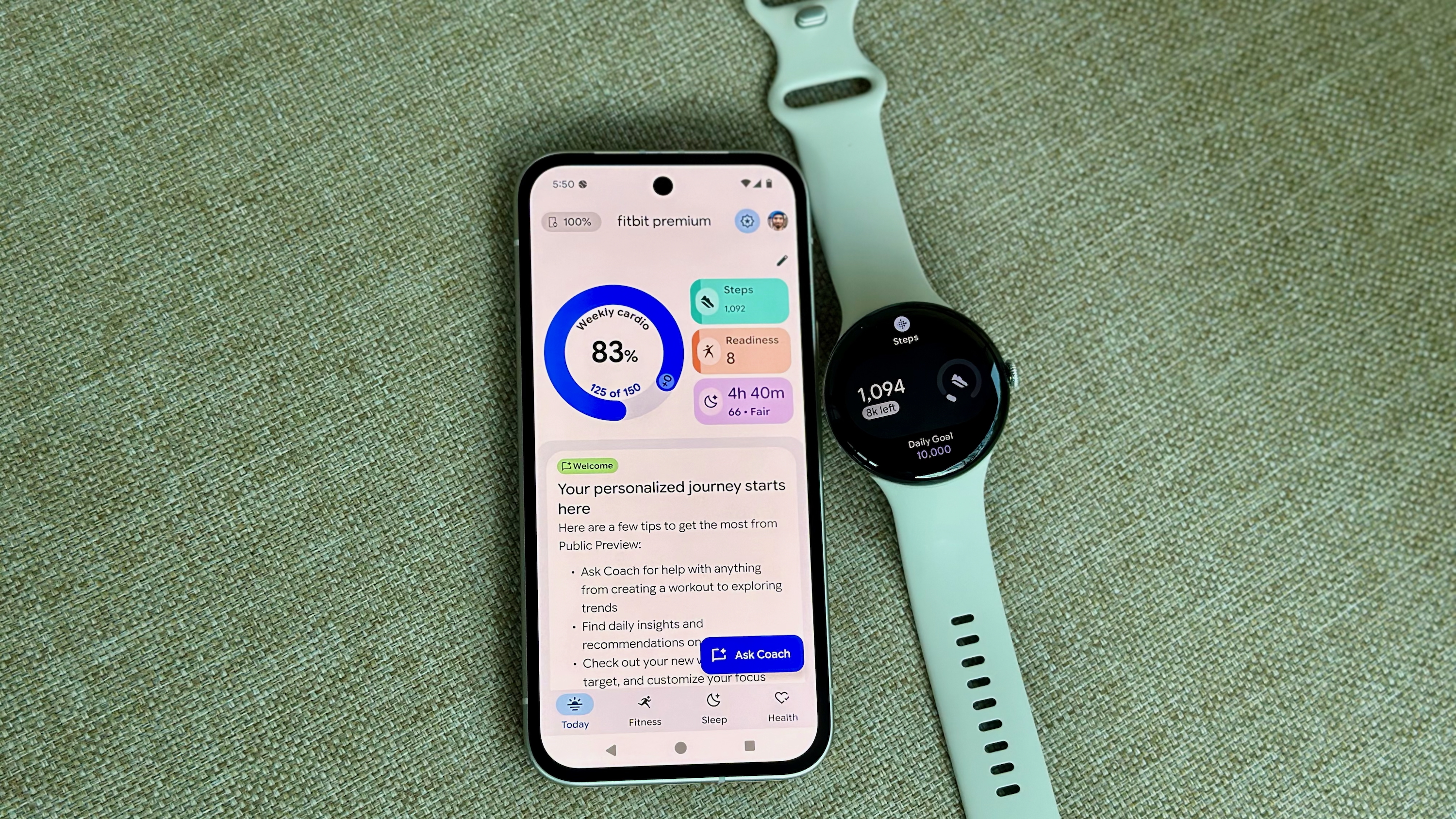 Μια φωτογραφία του Google Pixel Watch 4 που κάθεται δίπλα σε ένα Pixel 9a ανοιχτό στην εφαρμογή Fitbit, που δείχνει την προεπισκόπηση Fitbit Premium Health Coach με ανοιχτή την καρτέλα Σήμερα. εμφανίζει νέα γραφικά στοιχεία και πληροφορίες.