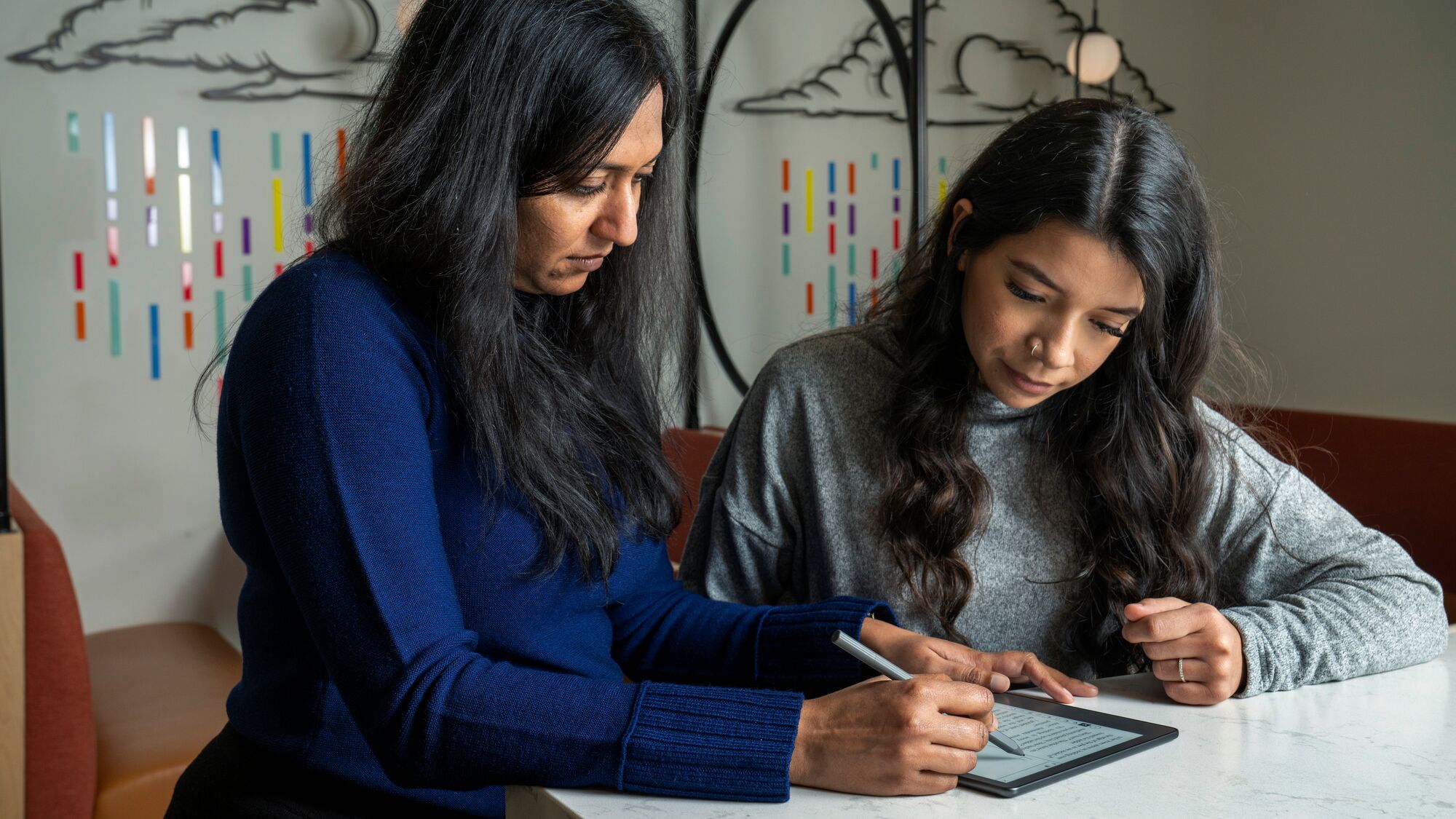 Two women focused on digital tablet at table with colorful wall art