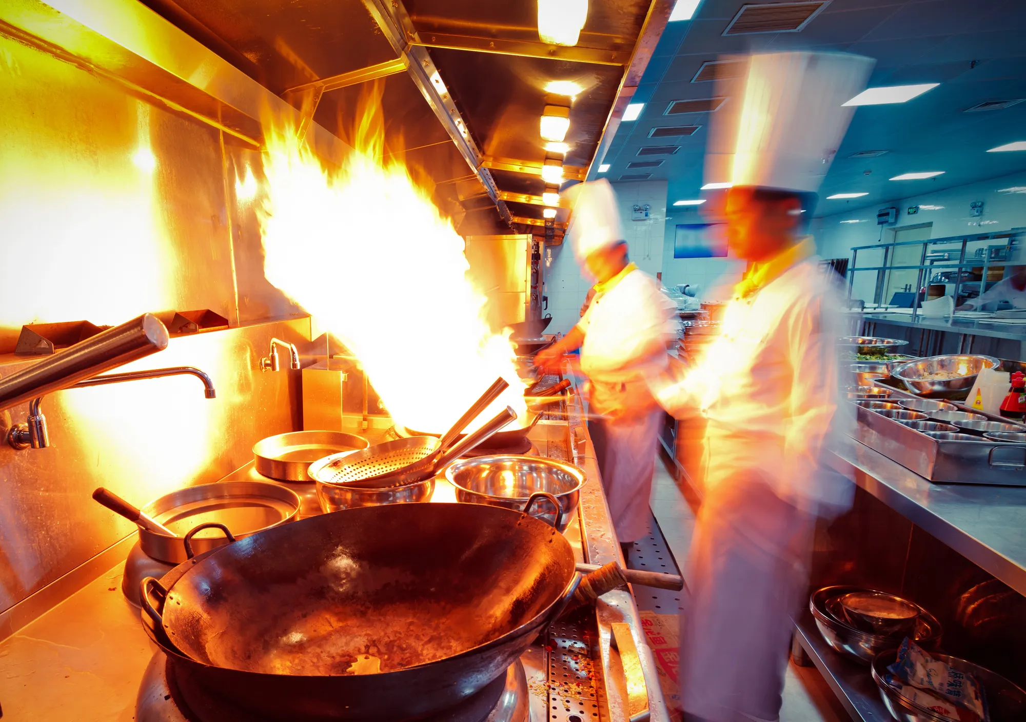 A wide-angle, action shot of a busy commercial kitchen with blurred motion, suggesting fast-paced cooking. Two chefs in white uniforms and tall hats are working at a stainless steel station. A large burst of flame erupts from a wok on a gas burner, and various cooking utensils and metal bowls are visible in the foreground.