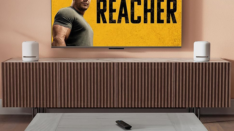 Ένα τηλεχειριστήριο Amazon Fire TV Stick σε ένα τραπέζι με μια τηλεόραση μπροστά του.