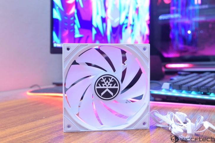 Ένας λευκός ανεμιστήρας InWin case με το λογότυπο της εταιρείας στο κέντρο είναι τοποθετημένος σε ξύλινη επιφάνεια, με πολύχρωμο στήσιμο gaming στο βάθος.