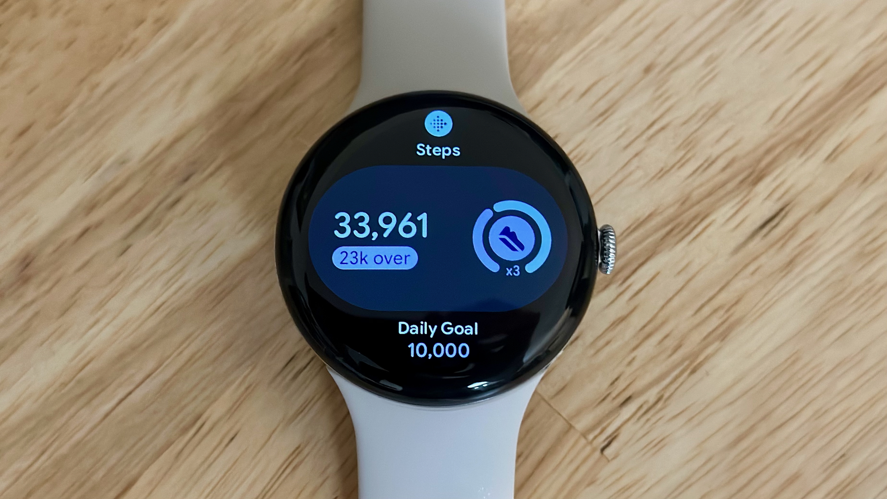 Μια φωτογραφία του Google Pixel Watch 4 σε ένα ανοιχτόχρωμο ξύλινο γραφείο που δείχνει το "Βήματα" Πλακίδιο, που δείχνει τρέχον αριθμό 33.961 με "Πάνω από 23 χιλιάδες" και από κάτω ημερήσιο στόχο 10.000.