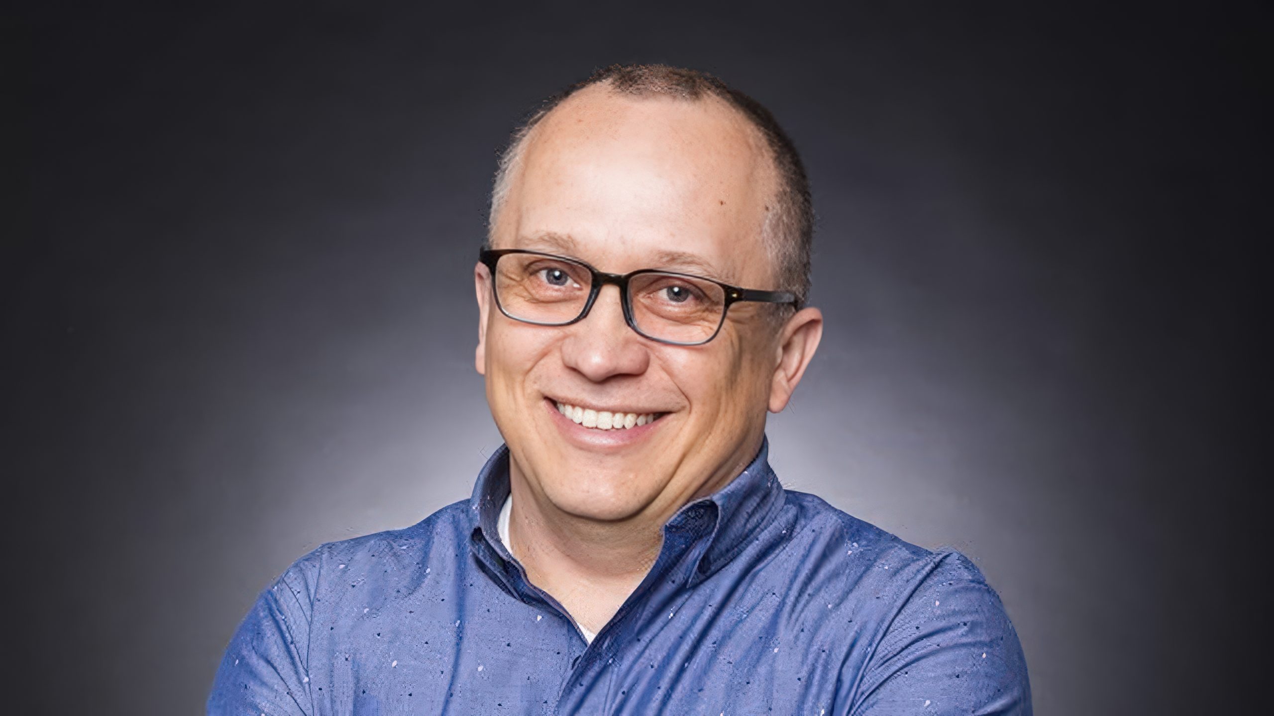 Is the MMO Genre Dead for Western AAA Developers? We Asked Three Industry Veterans A person wearing glasses and a blue patterned shirt, smiling against a dark gray background.