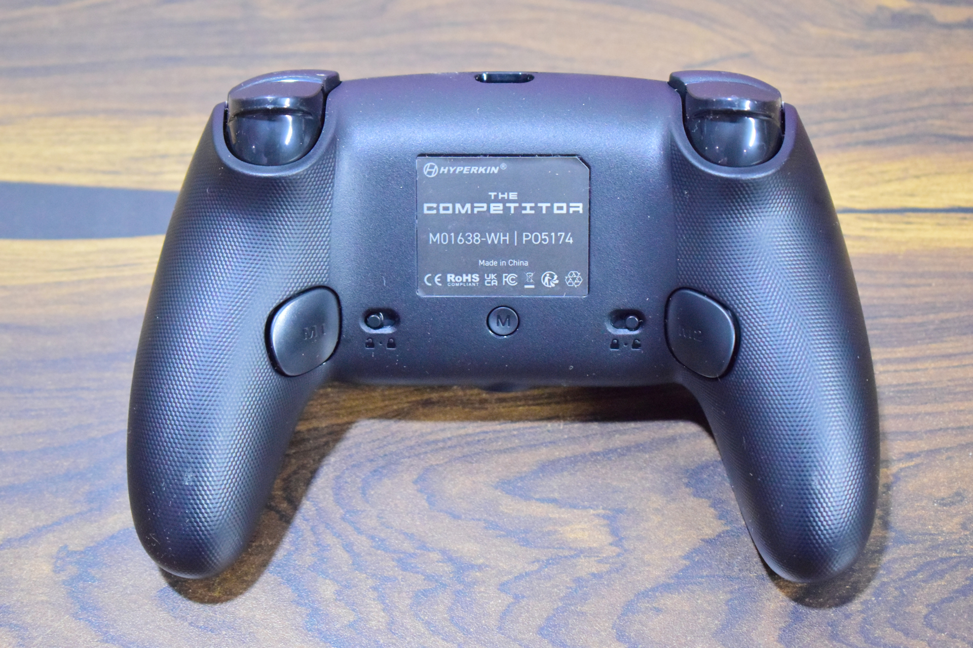 Η πίσω όψη ενός ελεγκτή Hyperkin The Competitor με αριθμό μοντέλου «M01638-WH» και «PO5174», που δείχνει διάφορα λογότυπα κανονισμών.