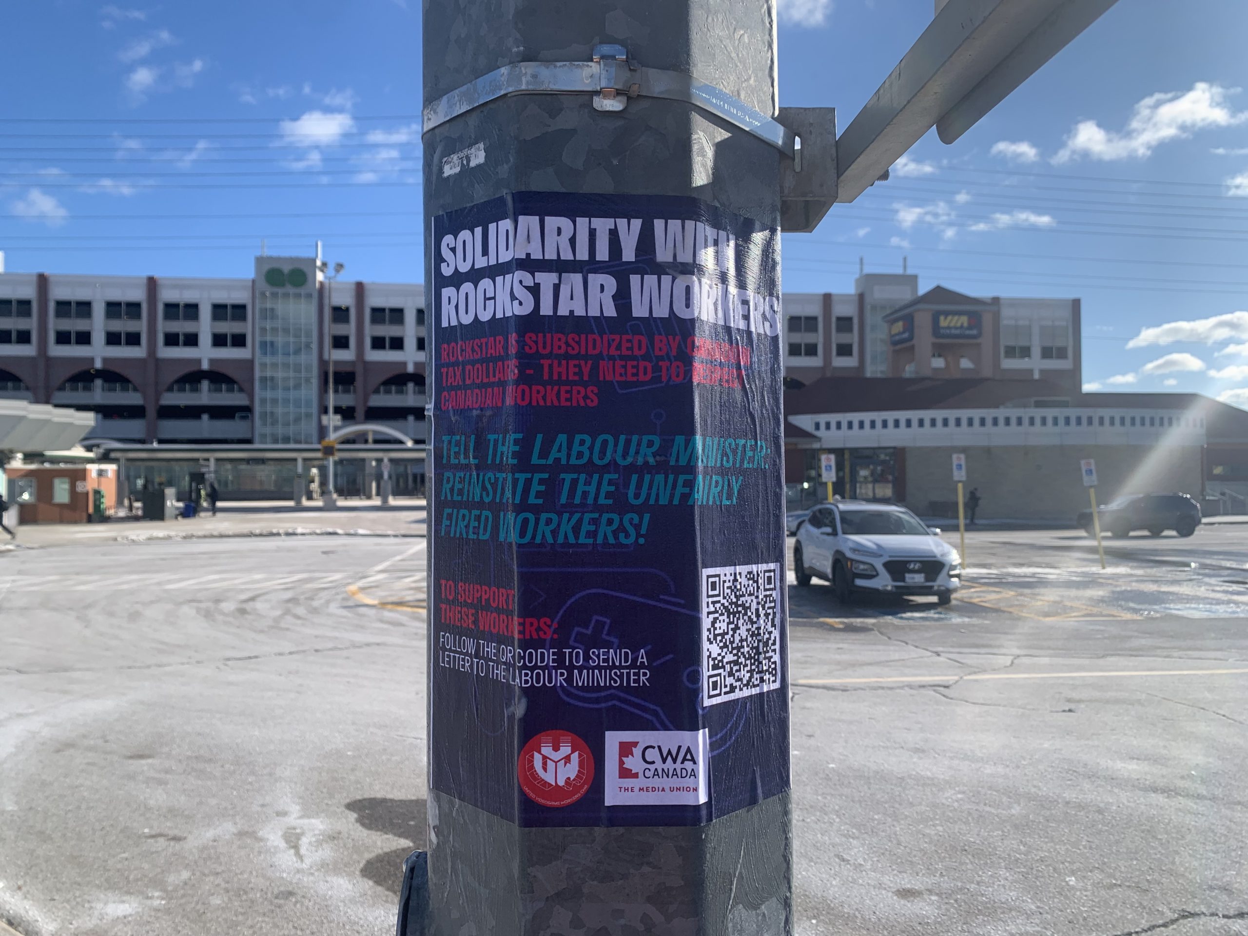 A poster on a pole in a parking lot reads 'Solidarity with Rockstar Workers,' urging the public to pressure the labor minister to reinstate fired workers, with a QR code and CWA Canada logo visible.