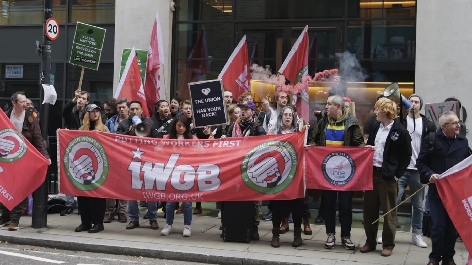 A group of people are holding flags and signs at a protest organized by the Independent Workers Union, with banners reading 'PUTTING WORKERS FIRST' and 'IWGB.ORG.UK'.