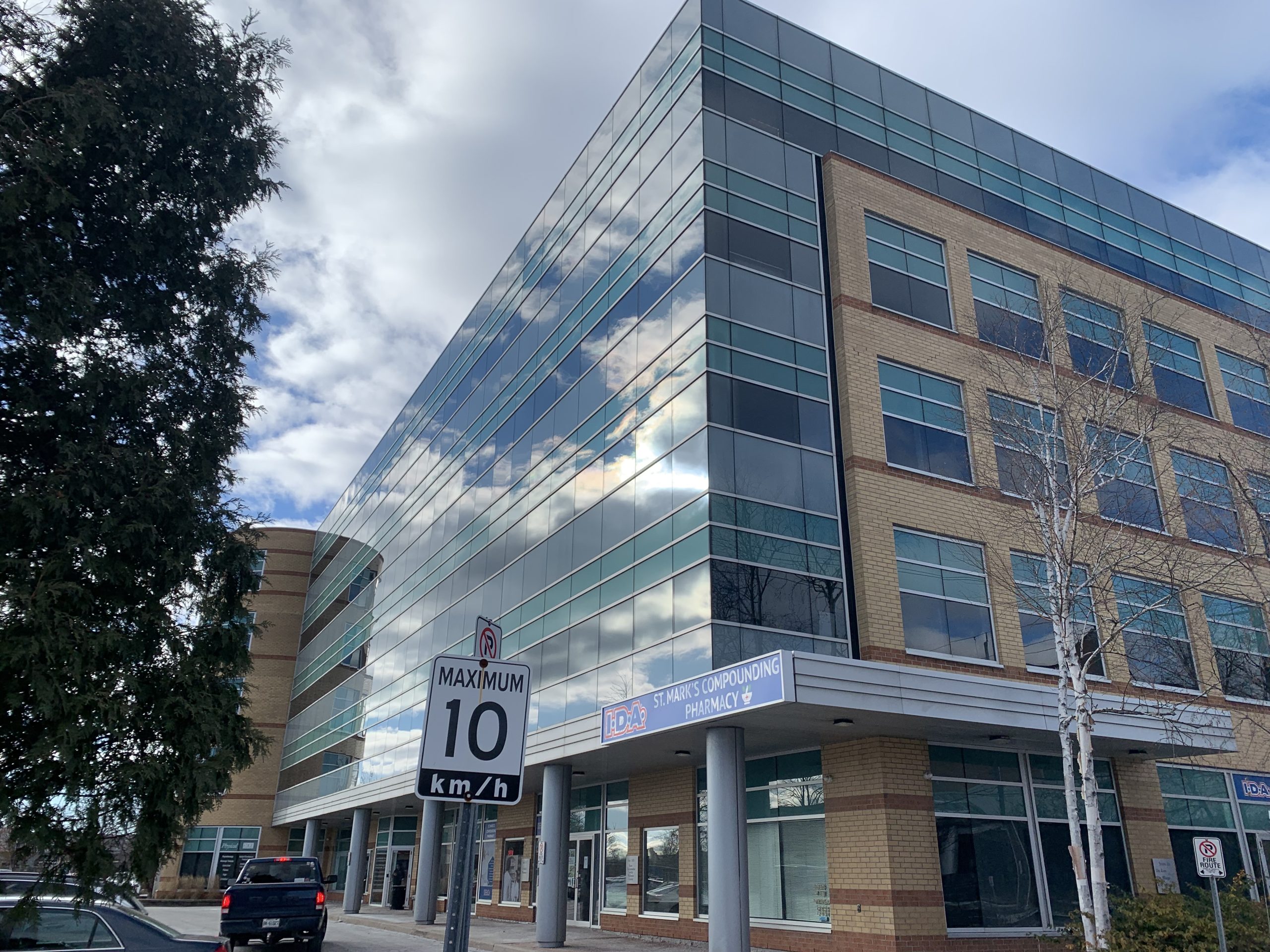 A multi-story office building with a sign reading 'St. Mark's Compounding Pharmacy' and a street sign displaying 'MAXIMUM 10 km/h'.