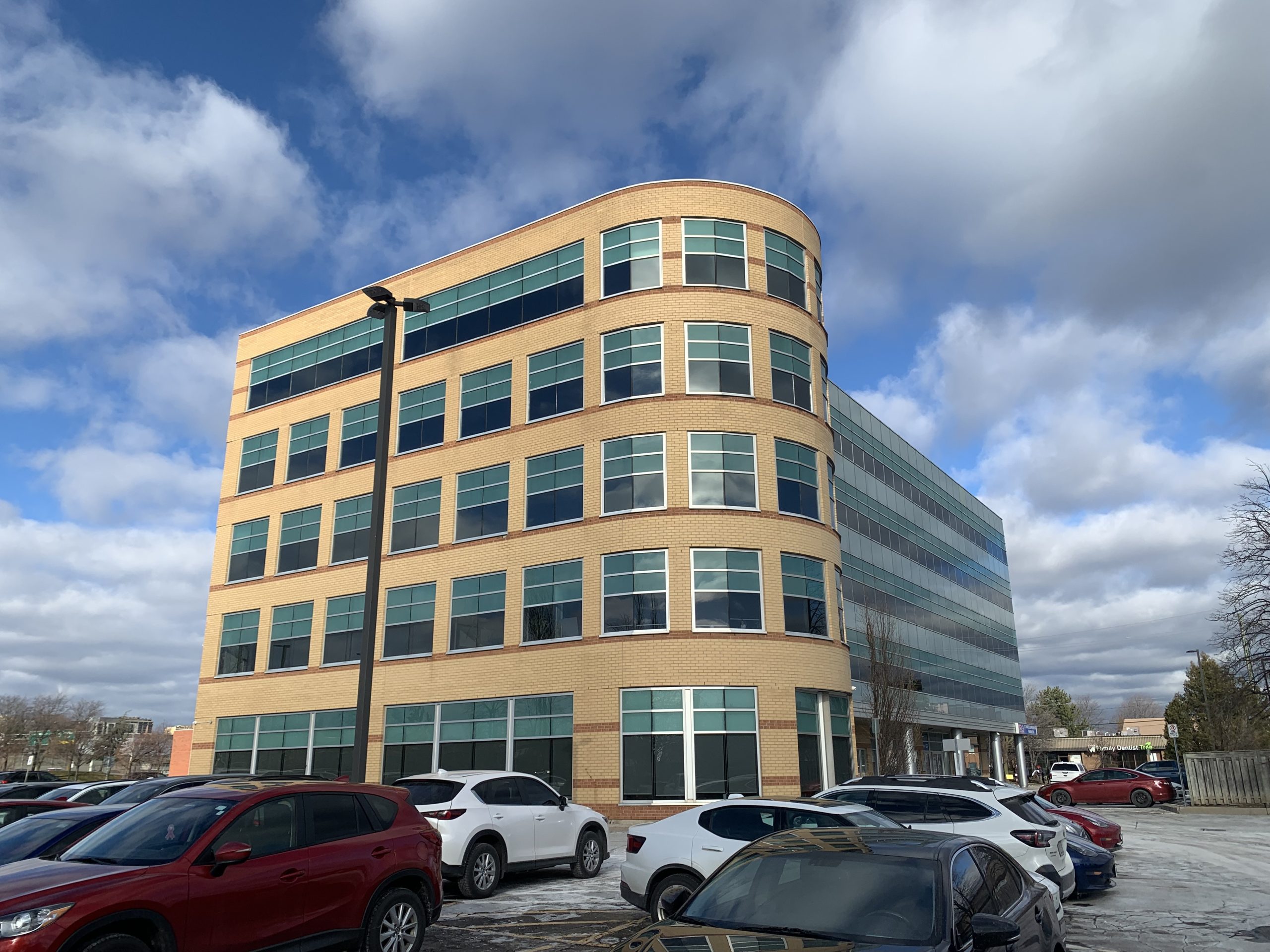 A multi-story office building with a rounded corner design and modern glass windows, surrounded by a parking lot with several parked cars.