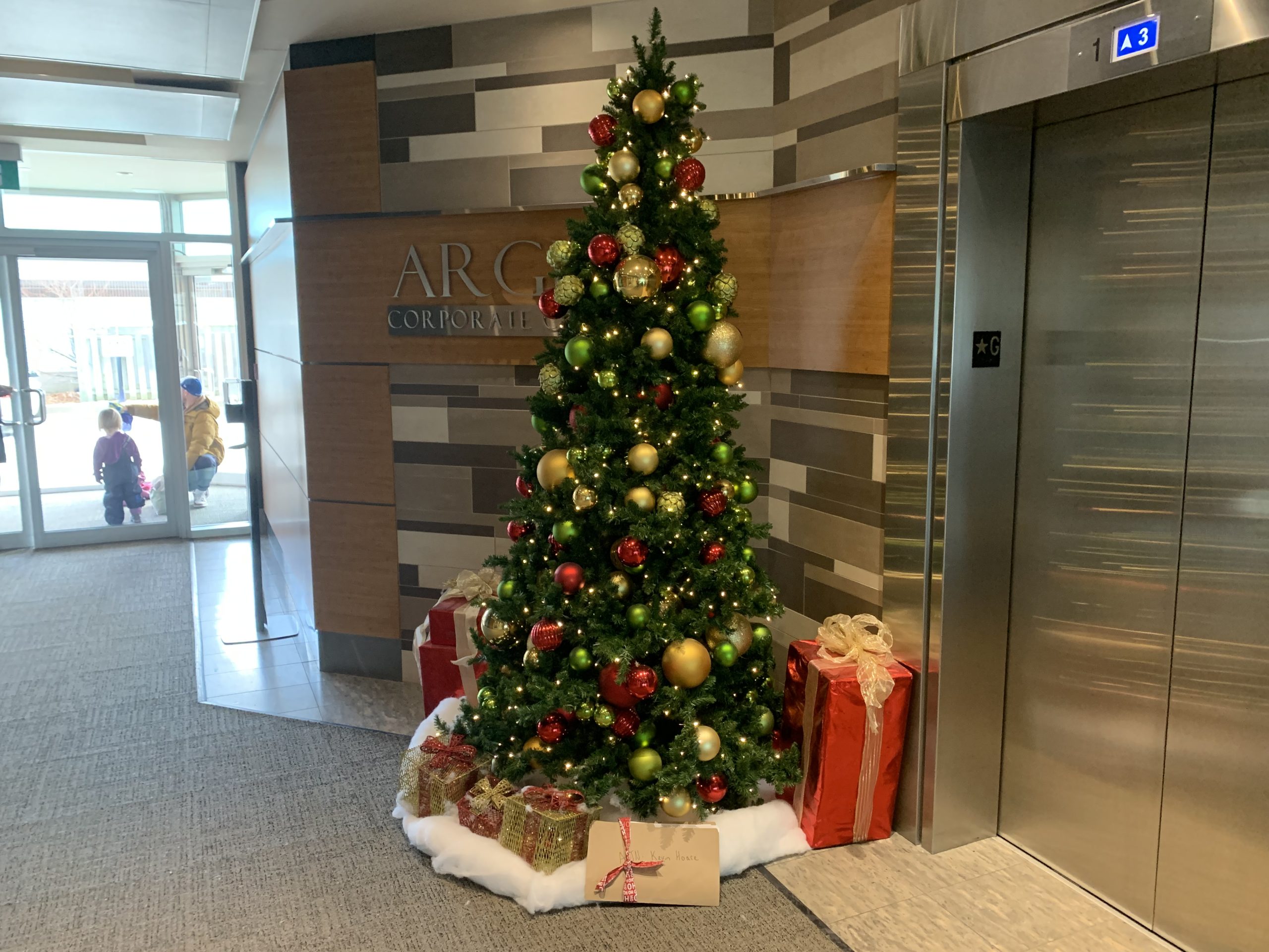 A decorated Christmas tree with gifts underneath stands next to an elevator in a corporate lobby labeled 'ARC.'