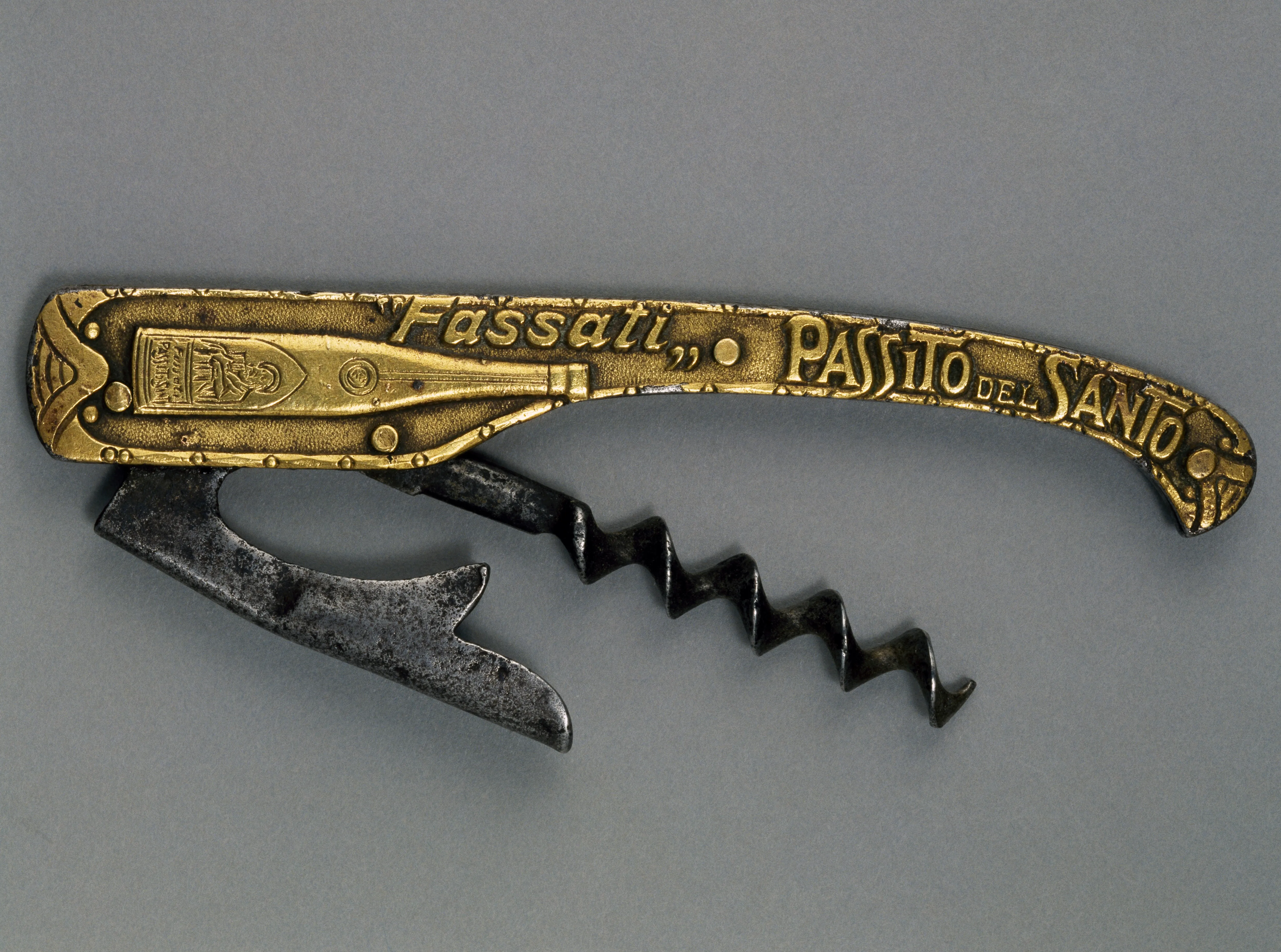 Ένα vintage πτυσσόμενο τιρμπουσόν σερβιτόρου (σχέδιο Carl Wienke) εμφανίζεται σε ουδέτερο γκρι φόντο. Η λαβή είναι κατασκευασμένη από ανάγλυφο μέταλλο σε χρυσό τόνο με το διακοσμητικό κείμενο "Fassati Passito del Santo" και μια απεικόνιση ενός μπουκαλιού κρασιού. Αναδιπλωμένο από τη λαβή είναι ένα σκούρο, μεταλλικό σπειροειδές σκουλήκι και ένας μοχλοβραχίονας με οδοντωτή ασημί τόνο, που δείχνει την πρώιμη μηχανική σχεδίαση του φορητού ανοίγματος κρασιού.