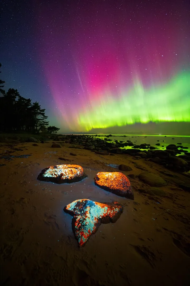 a pink and green aurora over a bitch with three rocks reflecting the glow