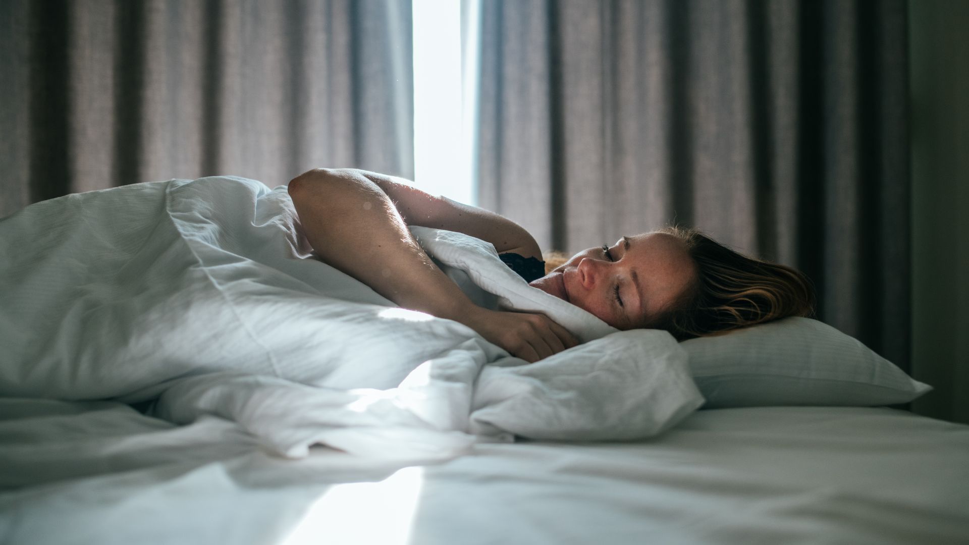 Μια γυναίκα κοιμάται σε ένα κρεβάτι που έχει ολόλευκα κλινοσκεπάσματα. Στο βάθος υπάρχουν κουρτίνες που είναι ελαφρώς ανοιχτές, αφήνοντας το φως του ήλιου να μπει.