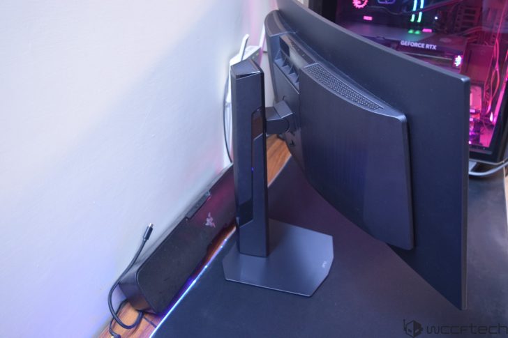 The image shows the back of a curved monitor on a stand next to a wall, with a Razer soundbar and gaming PC displaying a visible 'GeForce RTX' logo inside.