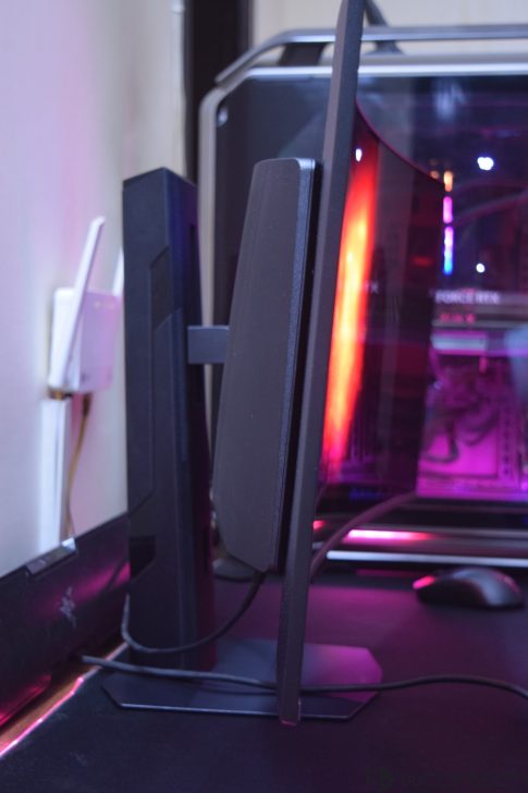 A side view of a black gaming monitor with a visible RGB-lit custom PC case in the background, featuring a 'GeForce RTX' logo inside.