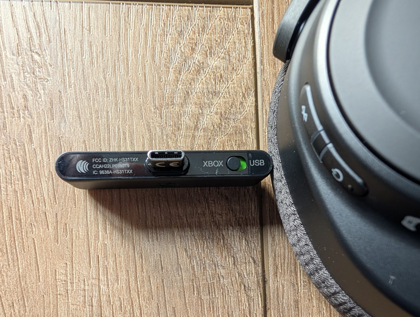 Ένα κοντινό πλάνο ενός ασύρματου προσαρμογέα USB Xbox με εμφανές αναγνωριστικό FCC «ZHK-HS31TXX» και άλλες σημάνσεις πιστοποίησης, δίπλα στο κουμπί λειτουργίας ενός ακουστικού.