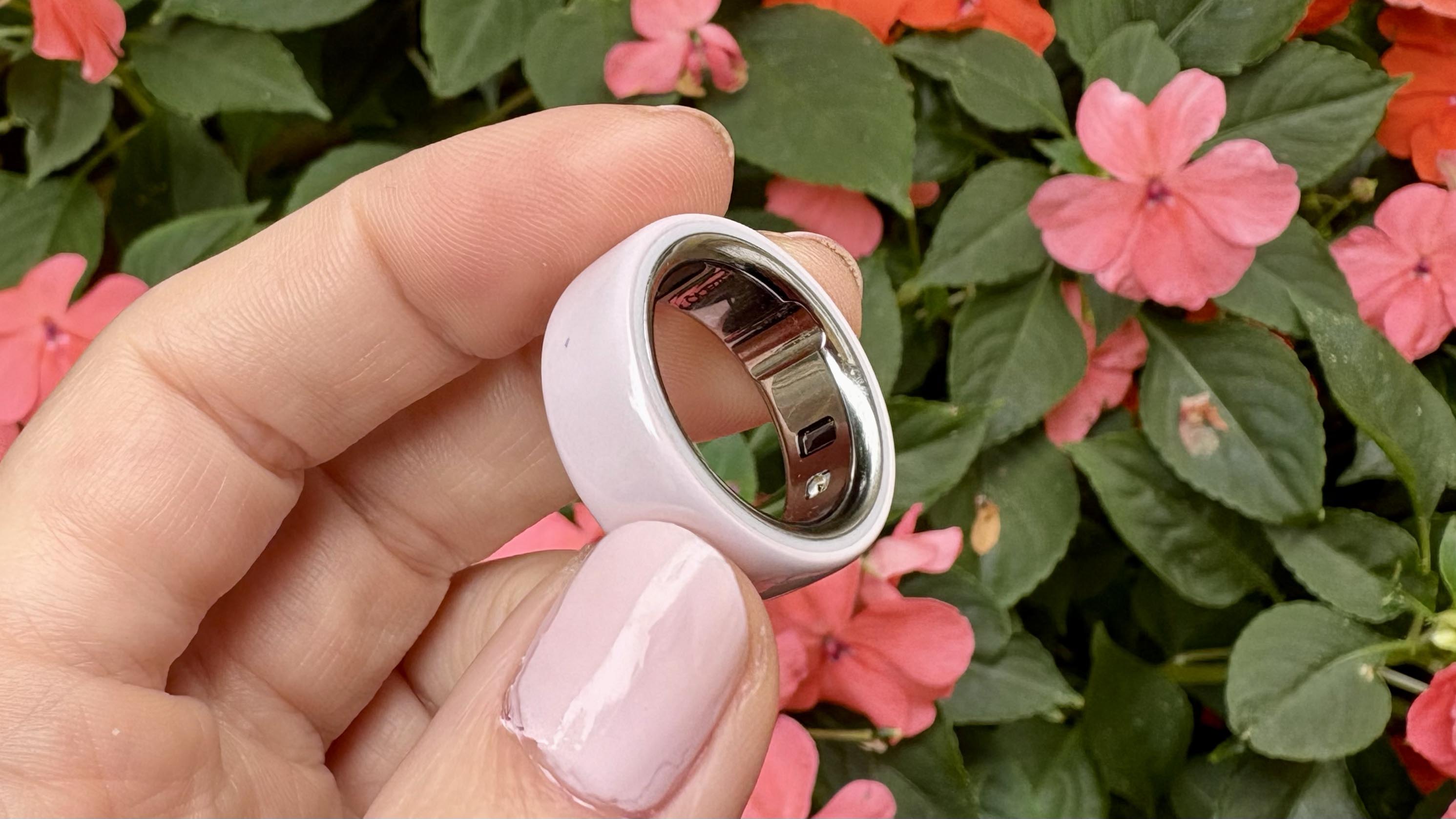 Το κεραμικό Oura Ring 4 ανάμεσα στα δάχτυλα ενός ατόμου μπροστά από μερικά λουλούδια