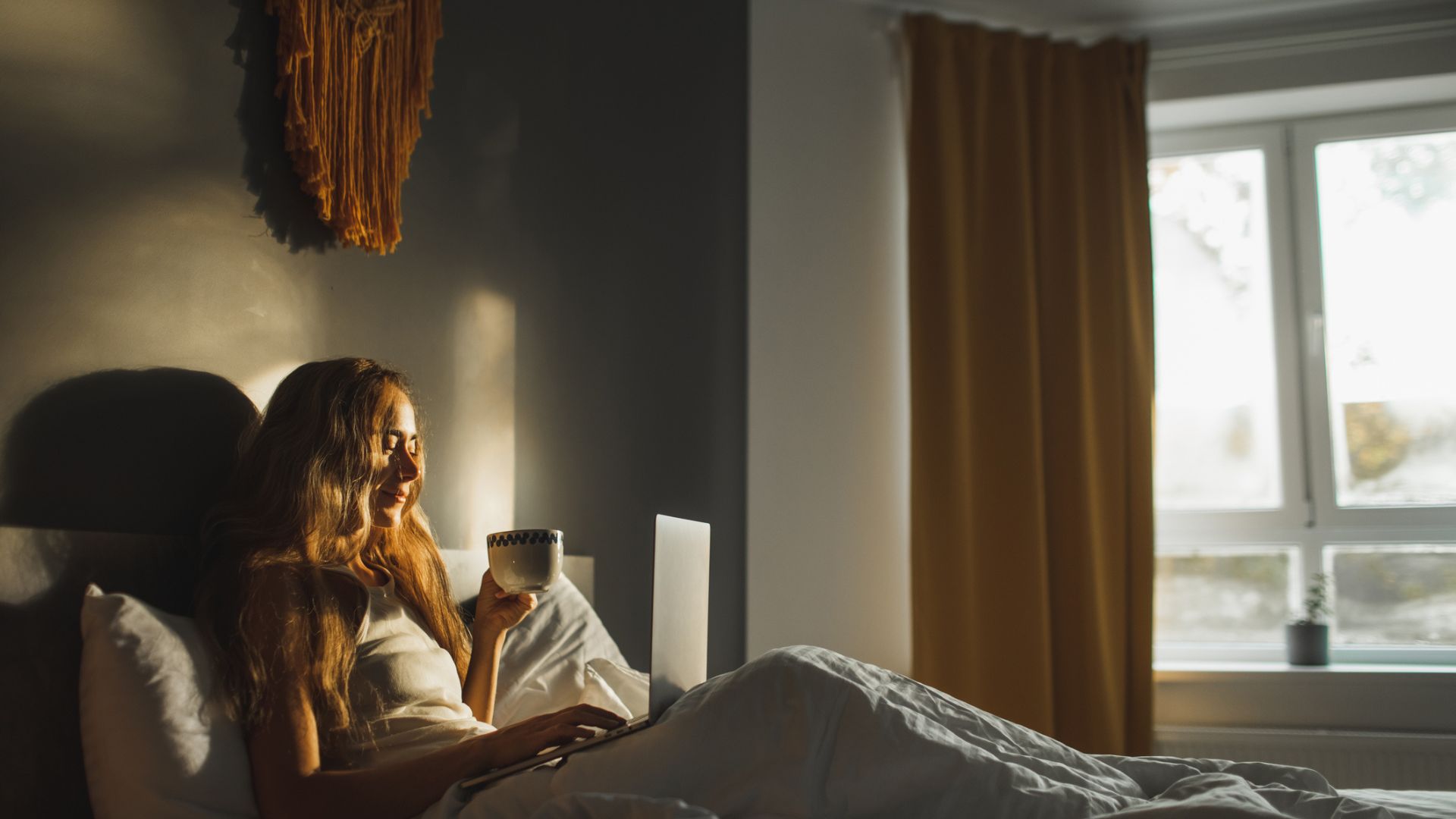 Μια γυναίκα κάθεται στο κρεβάτι πίνοντας ένα φλιτζάνι τσάι καθώς το φως του ήλιου περνάει μέσα από τις ανοιχτές κουρτίνες της