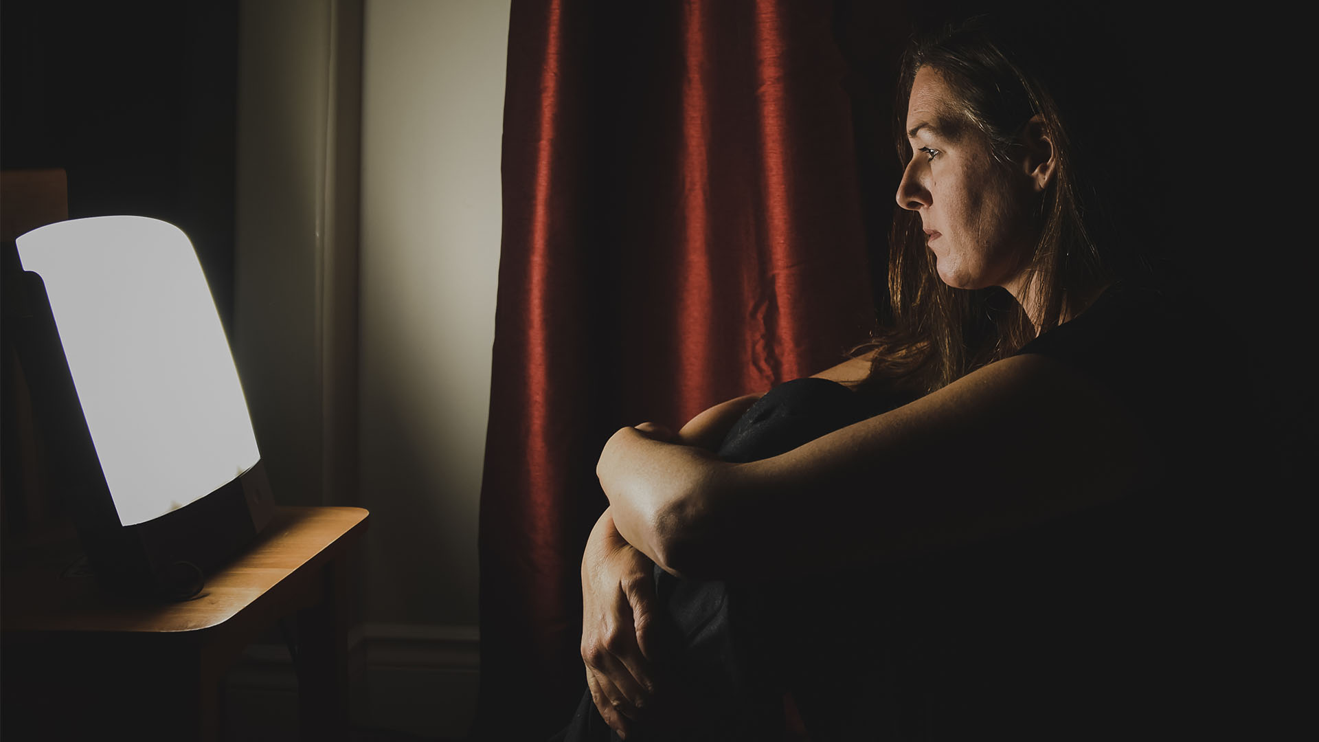 Μια γυναίκα κάθεται σε ένα σκοτεινό δωμάτιο μπροστά από τη φωτεινή λυχνία SAD φωτοθεραπείας της