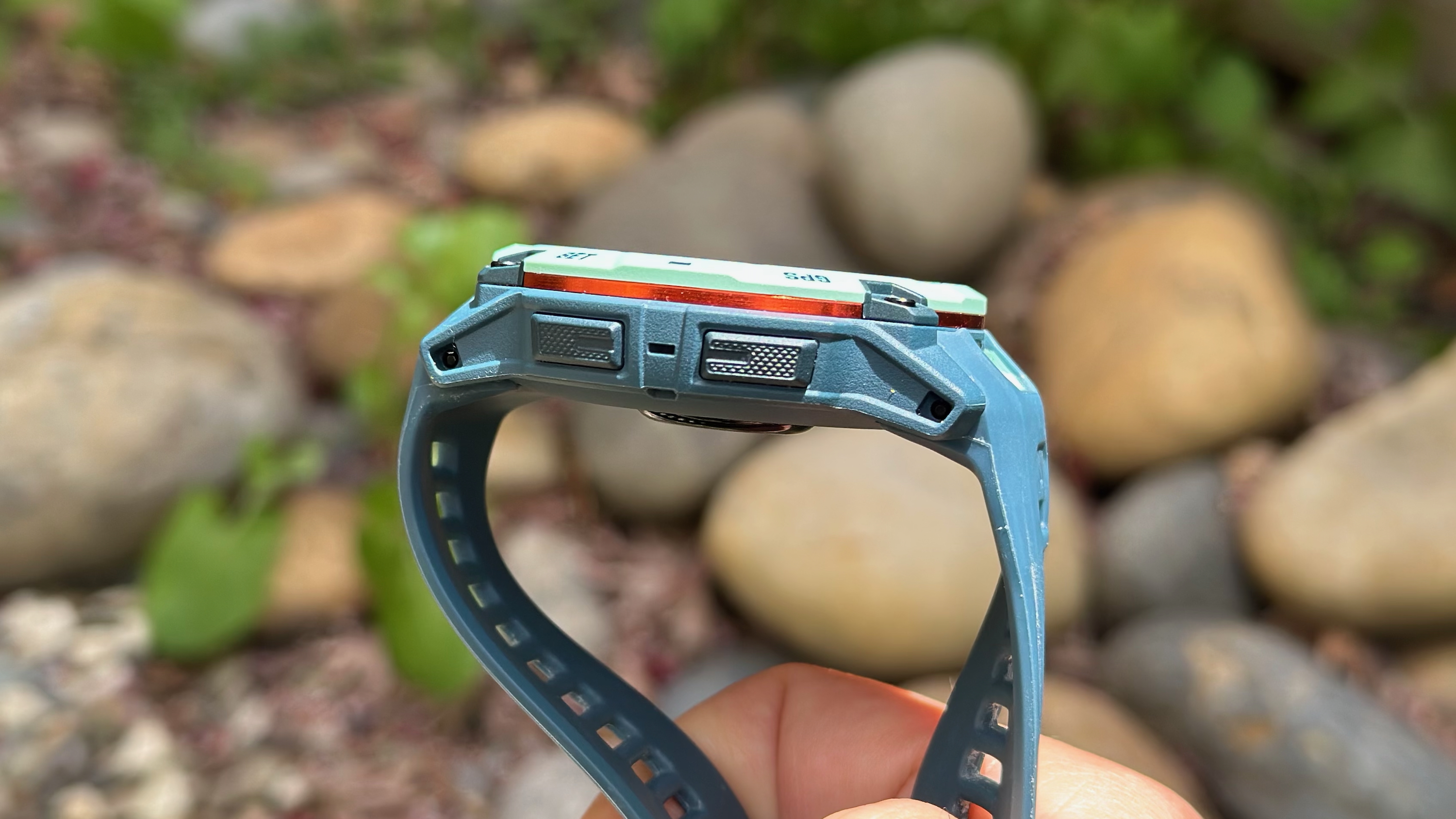 Η δεξιά πλευρά του Garmin Instinct 3 AMOLED 50mm, η φωτογραφία επικεντρώθηκε στα δύο κουμπιά και το πορτοκαλί αλουμινένιο πλαίσιο, με θαμπούς βράχους στο φόντο.