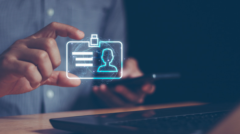 Ένα άτομο που κρατά μια εικονική εικονογράφηση ταυτότητας δίπλα στο τηλέφωνο και το φορητό υπολογιστή του