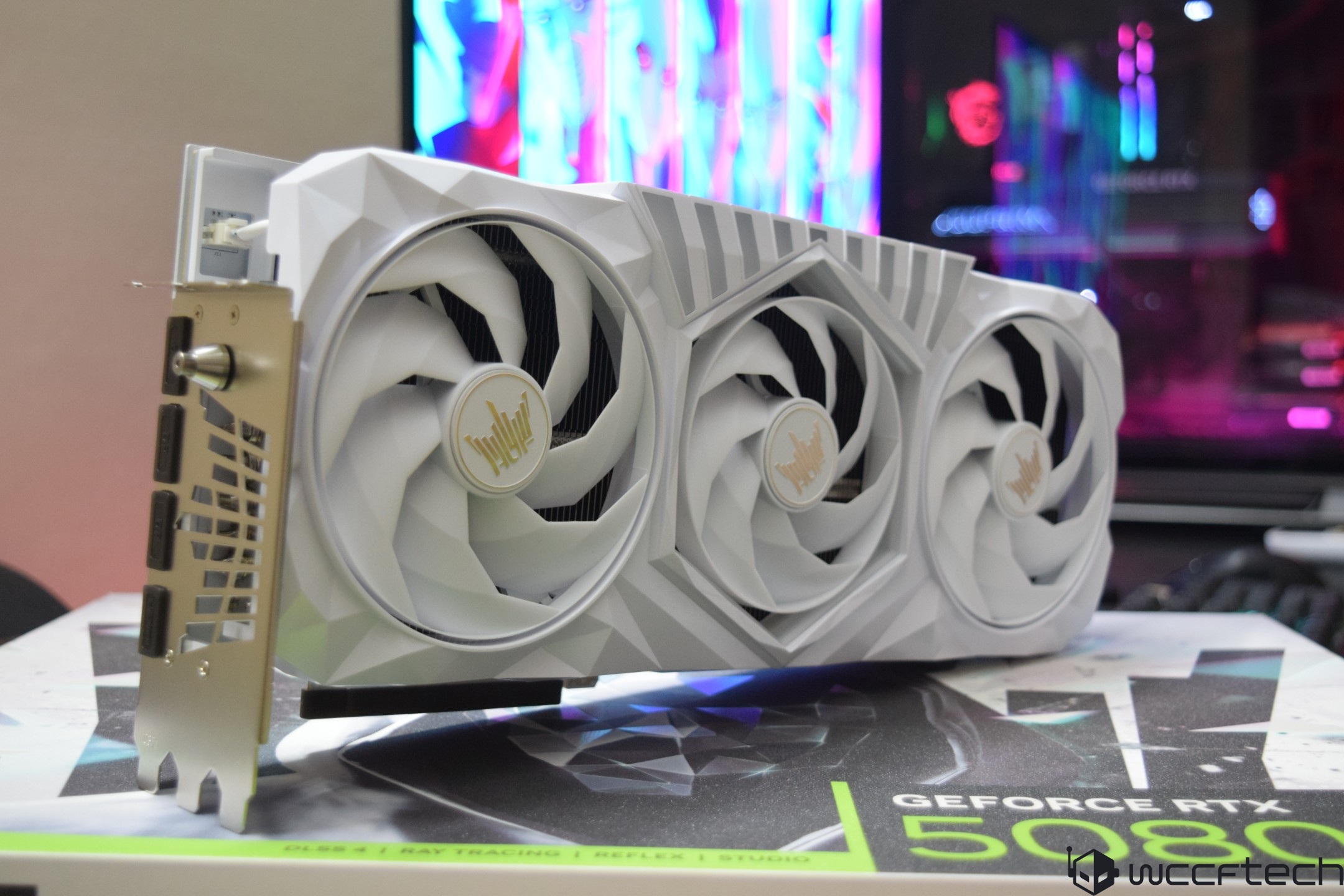 Μια λευκή κάρτα γραφικών με τριπλούς ανεμιστήρες βρίσκεται πάνω από ένα κουτί με την ένδειξη «GeForce RTX 5080 Ti DLSS 4» μπροστά από μια πολύχρωμη εγκατάσταση υπολογιστή.