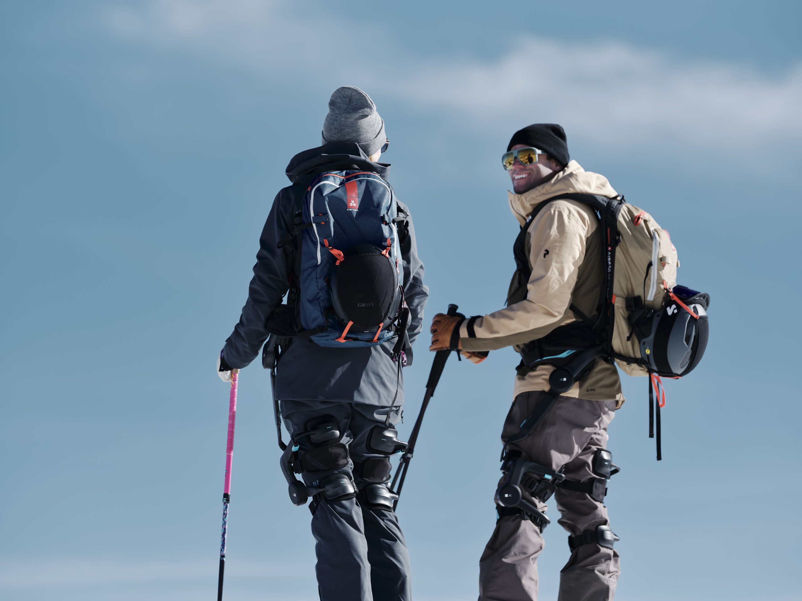 Δύο άτομα που φορούν κοστούμια και σακίδια από εξωσκελετό στέκονται με κοντάρια του σκι κάτω από έναν καταγάλανο ουρανό, ο ένας χαμογελά στην κάμερα.