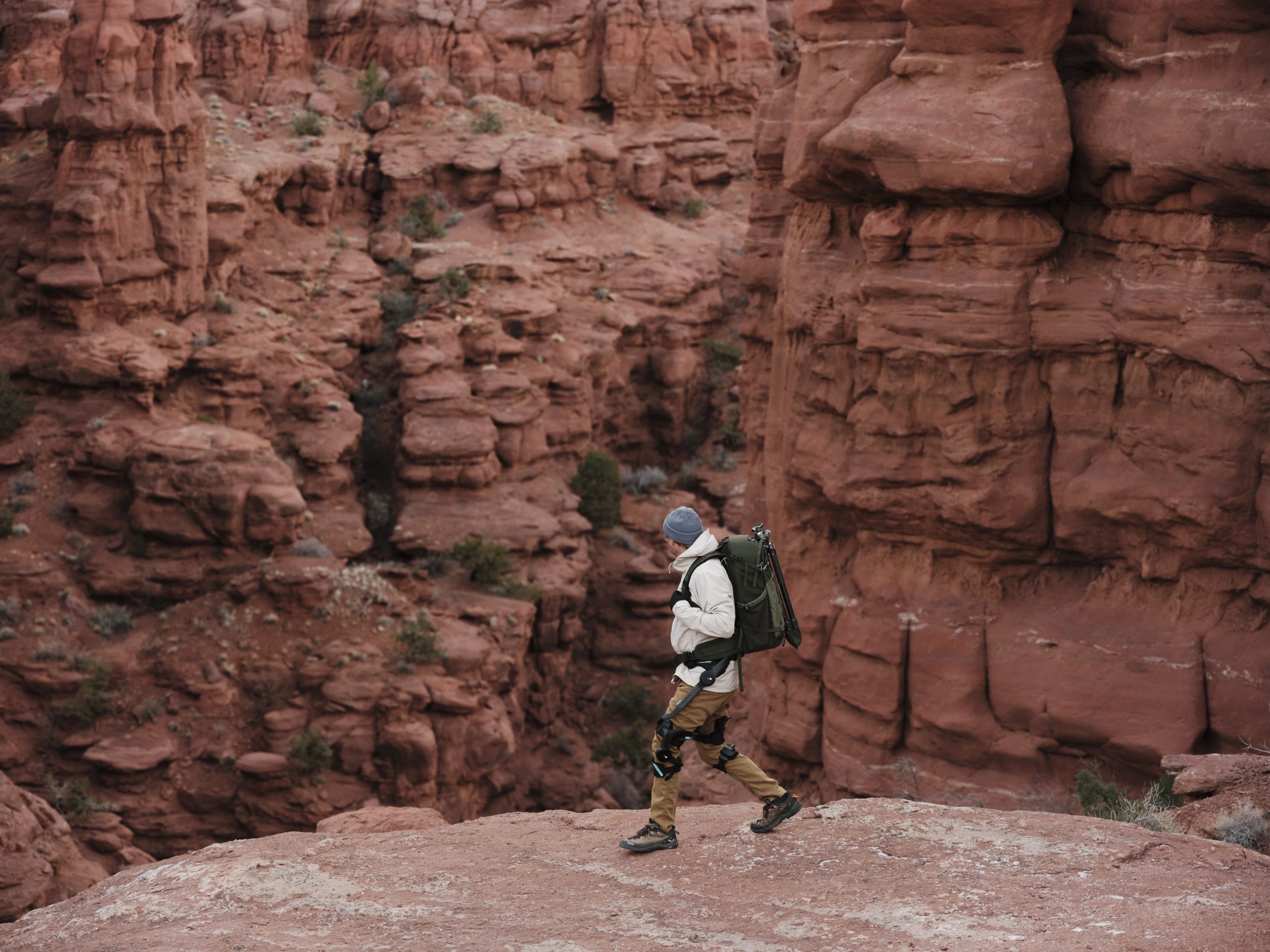 Ένα άτομο που φορά ένα σακίδιο και εξοπλισμό πεζοπορίας περπατά κατά μήκος ενός βραχώδους βράχου μέσα σε ένα κοκκινωπό τοπίο φαραγγιού.